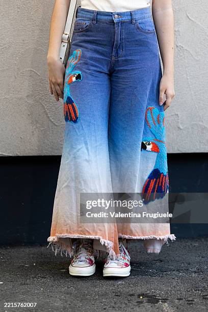 Guest wears blue and white embroidered tie-dye wide leg jeans, a white cross-body leather bag, a white t-shirt and printed sneakers at East Market...