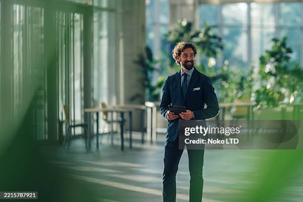 smiling businessman holding digital tablet walking in modern office - goed gekleed stockfoto's en -beelden
