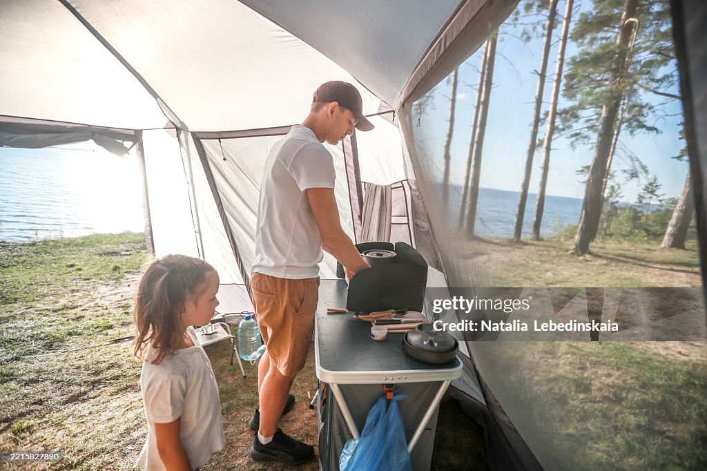 Summer Family camping: dad and his daughter is cooking breakfast on a camping stove in a tent under the morning sun together