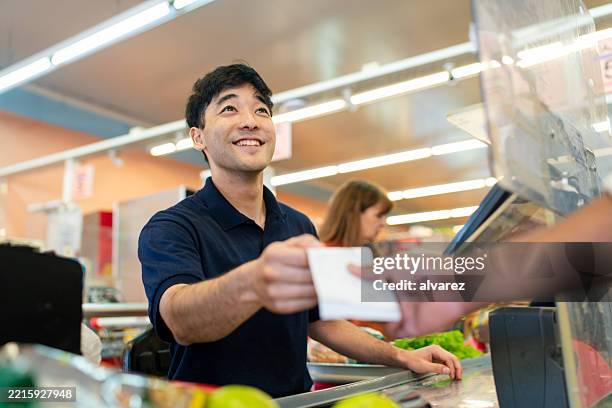 smiling cash register hands payment receipt to customer at supermarket checkout counter - grocery receipt stock pictures, royalty-free photos & images
