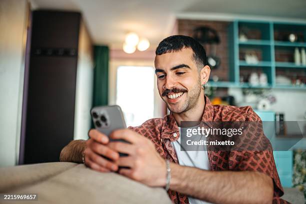 cheerful hipster man using smart phone while relaxing on a sofa at home - young men stock pictures, royalty-free photos & images