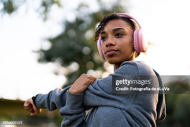 young woman stretching arms while listening music with headphones in a park - corredora de footing fotografías e imágenes de stock