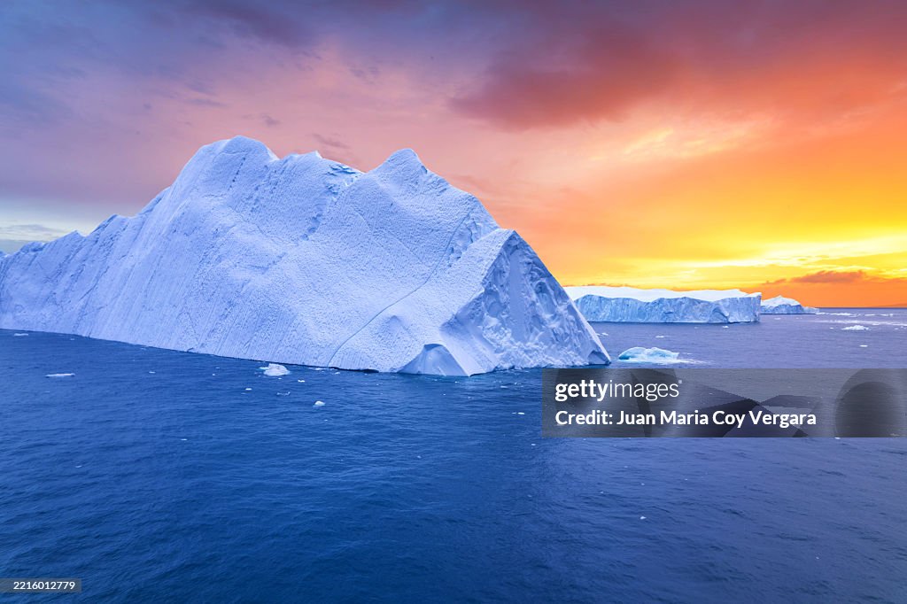 Aerial view of a massive iceberg floating silently in the icy waters of the Arctic Ocean near Ilulissat illuminated by a dramatic sky painted in vibrant shades of orange, yellow, and purple during the vibrant midnight sun. Ilulissat, Greenland