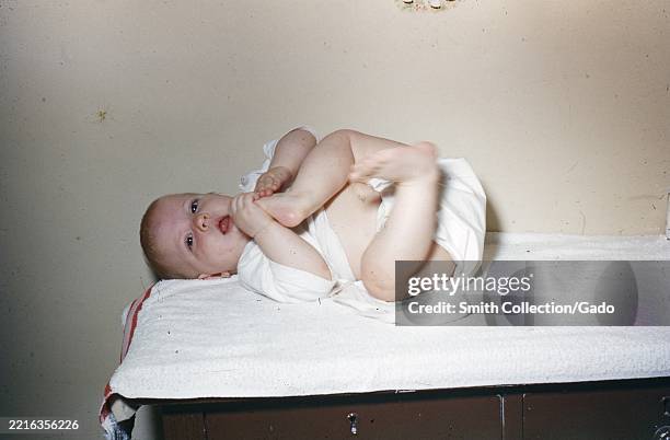 Infant in a white cloth diaper grasping a foot while lying on a terry-covered changing table inside a home, 1955.