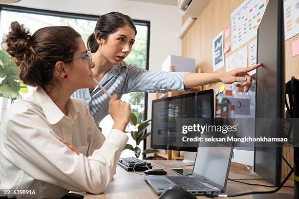 young female programmer is standing at a desk, writing code on her laptop, talking to her colleagues and giving advice. - software programmer stock pictures, royalty-free photos & images
