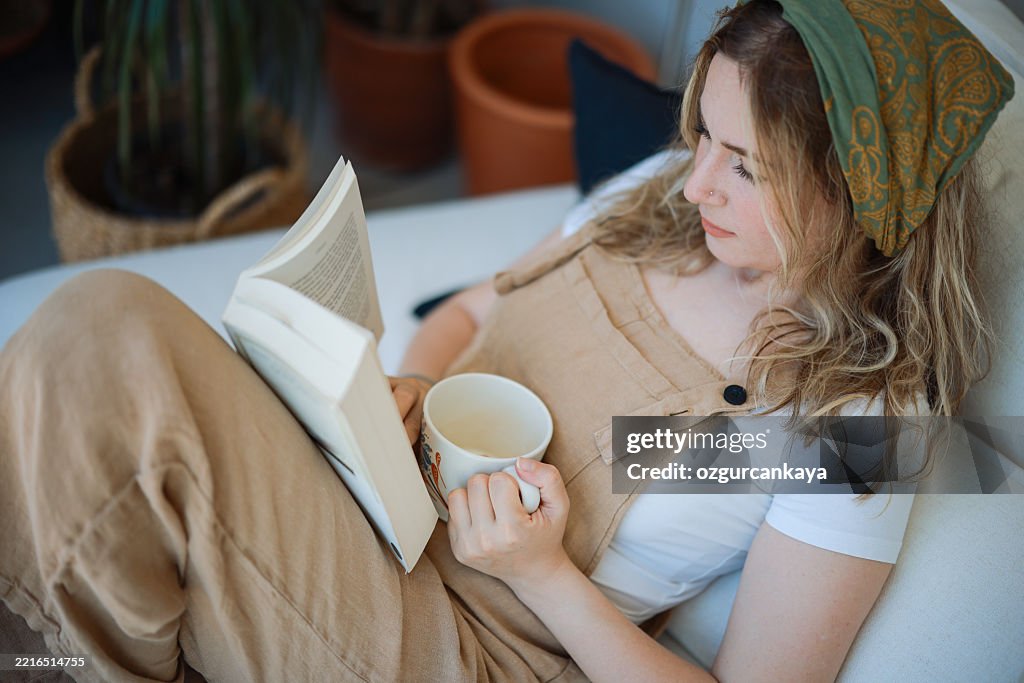 Young woman in overalls sitting on a sofa reading a book and drinking coffee