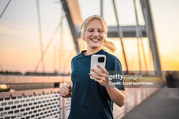 young sporty woman running and using her smartphone on a bridge at sunset - runner stock pictures, royalty-free photos & images