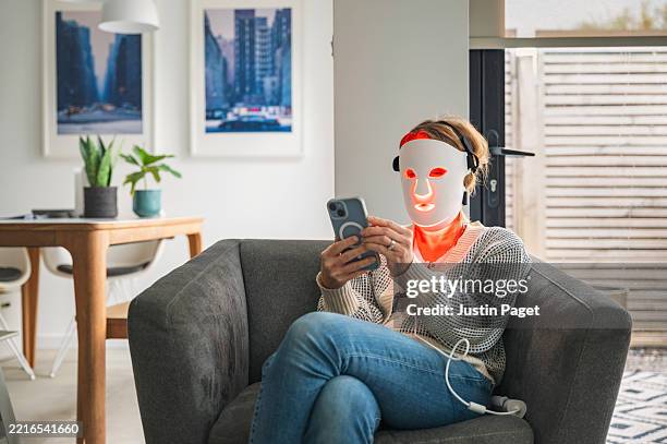 a mature woman uses her red led face mask (red light therapy) whilst relaxing at home in an armchair. she uses her smartphone - luce led foto e immagini stock