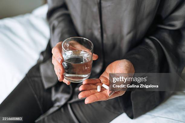 woman in pajamas drinking water with pills sitting on her bed at home - tomar remédio imagens e fotografias de stock