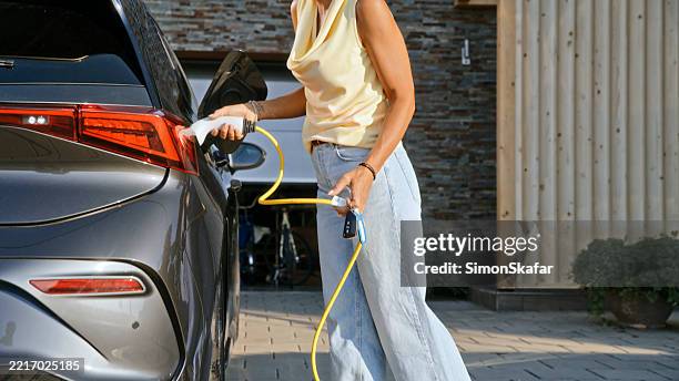 mid adult woman charging electric car in modern driveway on a sunny day - oprit stockfoto's en -beelden