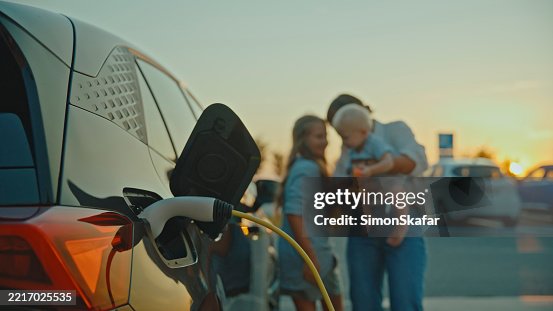 Caucasian Mother with Children Waiting Near Electric Car Charging at Sunset Caucasian Mother with Children Waiting Near Electric Car Charging at Sunset