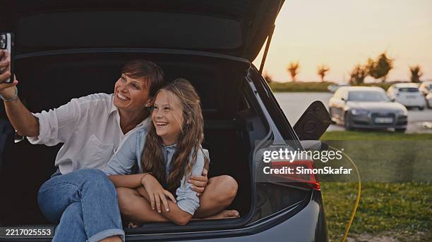 young mother and daughter taking selfie in electric car trunk during golden hour - plugging in stock pictures, royalty-free photos & images