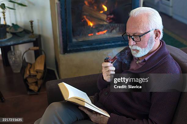 uomo anziano che legge il libro e fuma la pipa a casa - pipa prodotto a base di tabacco foto e immagini stock