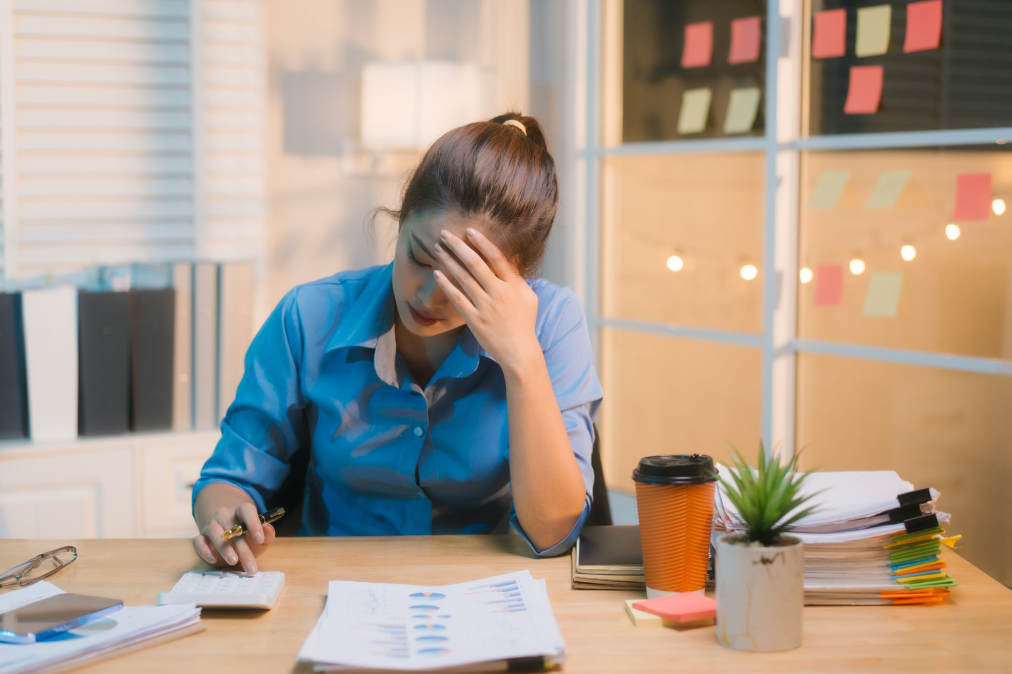 Working late into the night, Asian woman appears stressed and overwhelmed at her desk, rubbing her forehead as if experiencing migraine or headache, surrounded by paperwork and dimly lit office. Working late into the night, Asian woman appears stressed and overwhelmed at her desk, rubbing her forehead as if experiencing migraine or headache, surrounded by paperwork and dimly lit office.