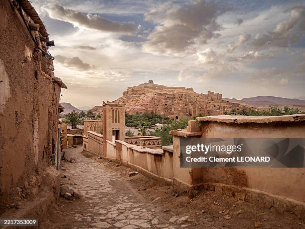 view of a cobblestone street lined with traditional houses and shops in the town of ait benhaddou at sunset, with the old berber fortified village and ksar (aït benhaddou kasbah) in the background, unesco world heritage site - ouarzazate - morocco. - ait benhaddou stock-fotos und bilder