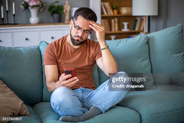 joven estresado leyendo malas noticias en el teléfono móvil en casa - enredado fotografías e imágenes de stock