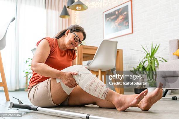 eine reife frau mit bandagiertem bein fiel zu hause zu boden. sie berührt ihr knie mit schmerzverzerrtem gesicht - gebrochenes bein stock-fotos und bilder