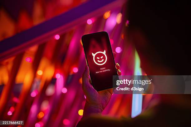 female hand holding smartphone showing an evil with devil horn on her ai chatbot conversation app in the city. illustrating the ideas around the risks and dangers of artificial intelligence (ai). the awareness and careful consideration of its implications - risque photos et images de collection