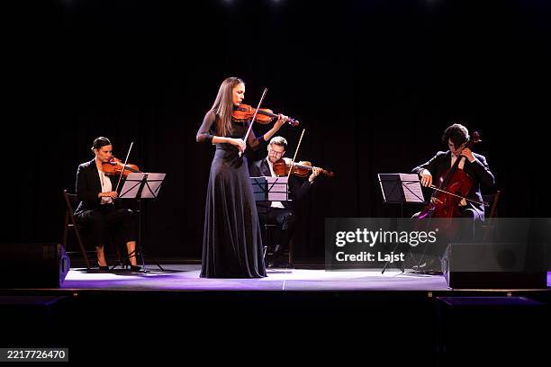 elegante violinista con un elegante vestido negro tocando el violín mientras está de pie frente al resto del cuarteto de cuerdas - clásico fotografías e imágenes de stock