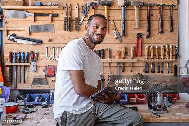 confident mid-adult carpenter using digital tablet in well-organized workshop with tool wall - handyman stock pictures, royalty-free photos & images