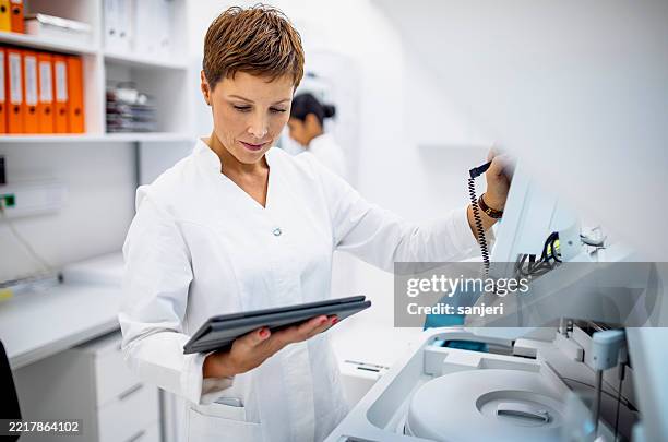 wissenschaftlerin im mittleren erwachsenenalter, die ein digitales tablet verwendet, während sie laborgeräte in einer modernen forschungseinrichtung bedient - krankenblatt stock-fotos und bilder