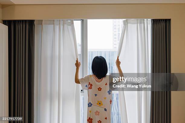 a young asian woman, an early riser, stands by the window and pulls back the white curtains to look out at the cityscape - look back at early colour photography stockfoto's en -beelden