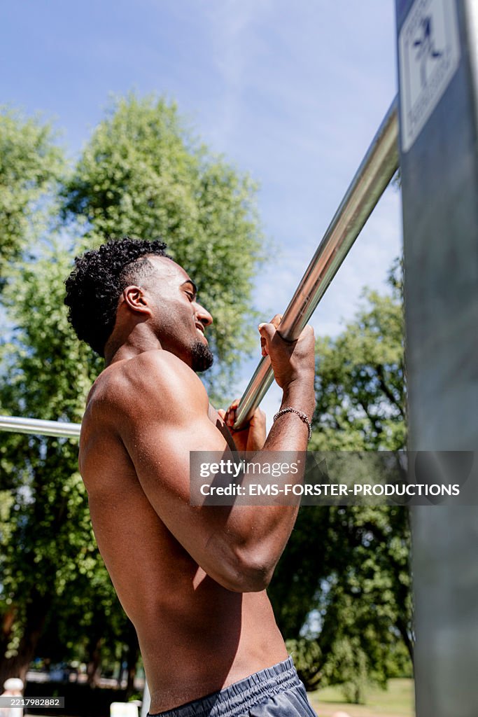 Sporty muscular build young black man is doing strength training outdoors with pull-ups on a metal bar in a publik park while sunny Sommer day.