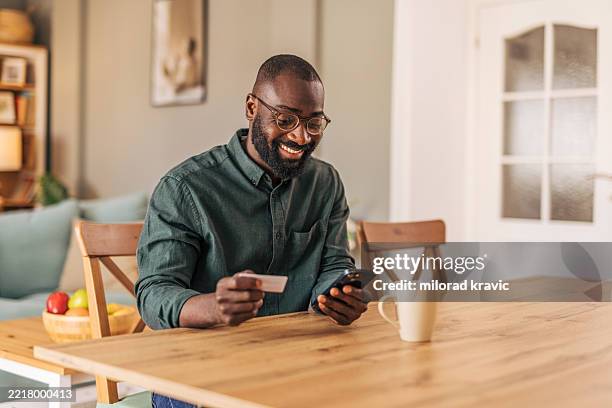smiling man making online payment using credit card and smartphone at home - spending money stock pictures, royalty-free photos & images