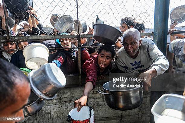 Palestinians stretch their arms through a metal fence with empty pots and pans, trying to receive food from a charity distribution point in Khan...