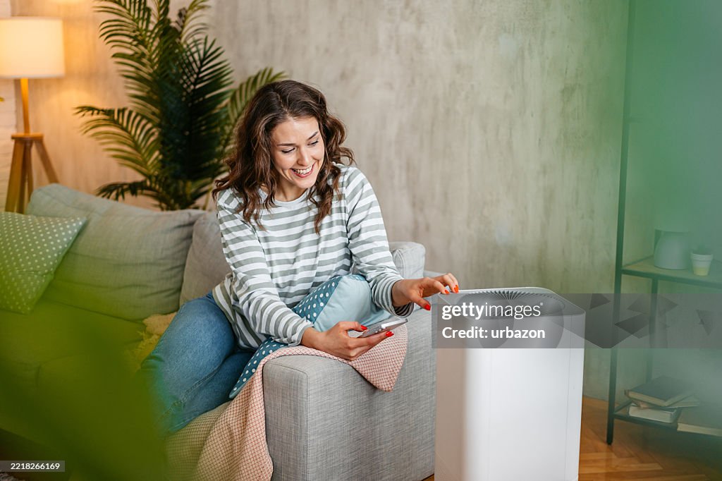 Young Woman Turing On The Air Purifier Using Her Phone While Sitting On The Sofa At Home