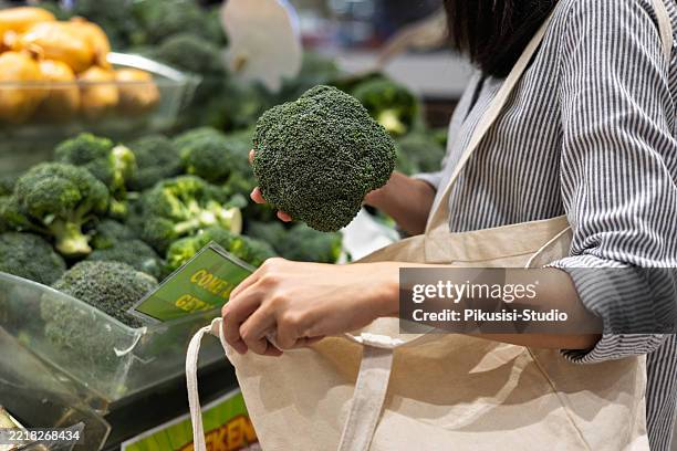 woman choosing fresh broccoli at supermarket with reusable bag - herbruikbare tas stockfoto's en -beelden