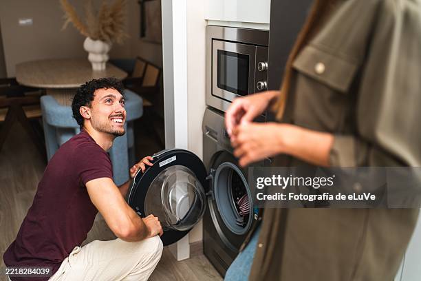 young man loading clothes into washing machine and looking at his partner - tarea doméstica fotografías e imágenes de stock