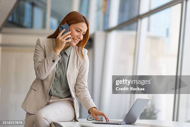 professional businesswoman working on laptop, talking on smartphone while seated at desk in contemporary workplace - saleswoman stock pictures, royalty-free photos & images