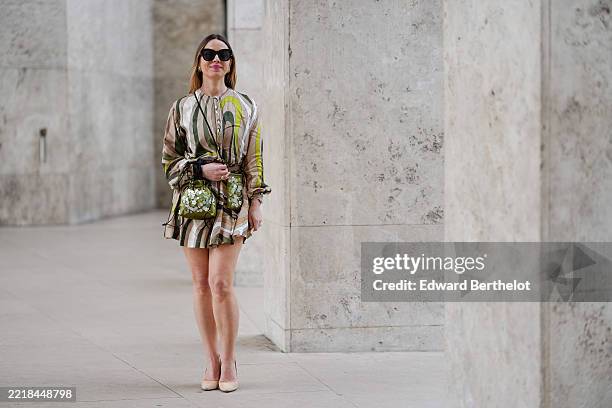 Maria Rosaria Rizzo wears oversized black sunglasses and gold hoop earrings. The hair is styled straight and loose. The dress features a multicolored...