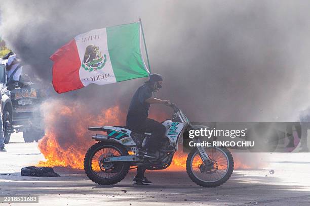 Demonstrator waves a Mexican national flag next to a car on fire during a protest following federal immigration operations, in the Compton...
