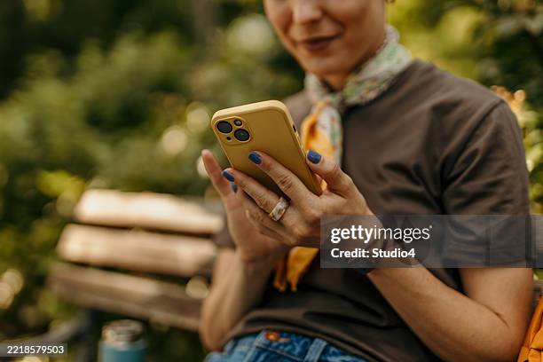 woman using smartphone while relaxing on park bench - vasthouden stockfoto's en -beelden