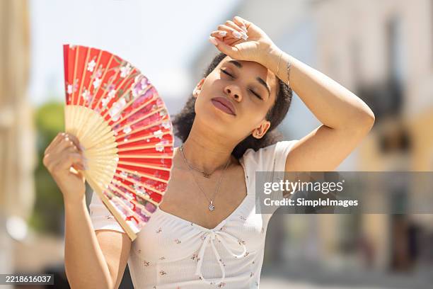 portrait of a young woman sitting in hot weather bench in city, cooling herself with a hand fan and wiping sweat from her forehead. - hand fan stock pictures, royalty-free photos & images