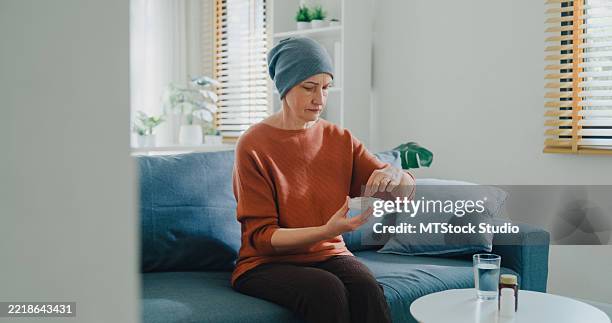 seniorin, krebspatientin mit kopftuch, die verschreibungspflichtige pillen mit wasser einnimmt, sitzt auf dem sofa im wohnzimmer zu hause. leben mit krebs konzept. - tabletten schlucken stock-fotos und bilder