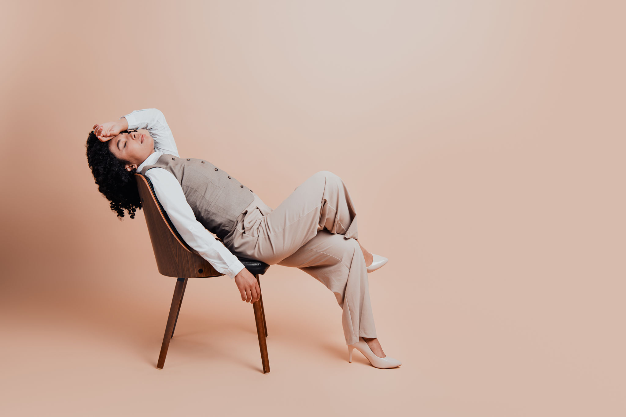 Professional woman wearing formal attire sitting on a wooden chair in a stylish pose against a beige background Professional woman wearing formal attire sitting on a wooden chair in a stylish pose against a beige background