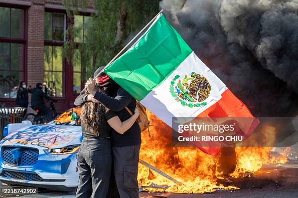 Waymo Flag TOPSHOT - A couple with a flag embraces as a Waymo vehicle burns, as ...