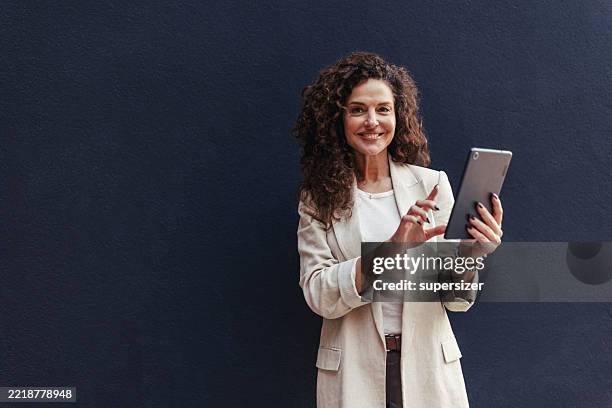 mujer segura de sí misma que sostiene la tableta contra la pared oscura - una mujer de mediana edad solamente fotografías e imágenes de stock