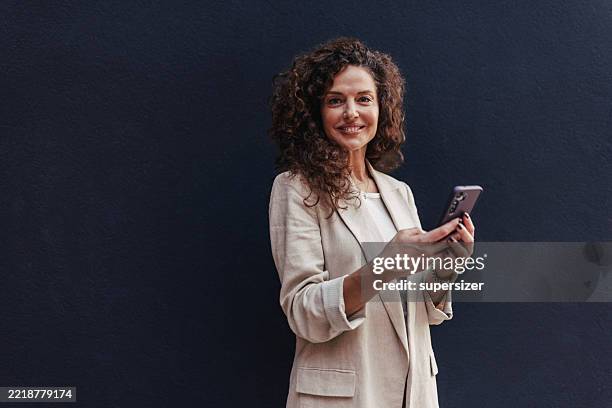 mujer sonriente en un atuendo elegante con teléfono inteligente al aire libre - una mujer de mediana edad solamente fotografías e imágenes de stock
