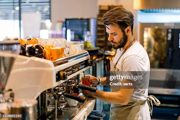 young male barista preparing espresso with a professional coffee machine in a stylish modern café interior - barista stockfoto's en -beelden