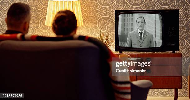 mid-adult couple watching vintage television program in warmly lit living room with retro decor - old watch stockfoto's en -beelden