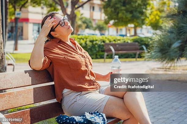 displeased woman eyes closed sitting on a park bench holding a water bottle. suffering from heat stroke and headache - temperatuur stockfoto's en -beelden