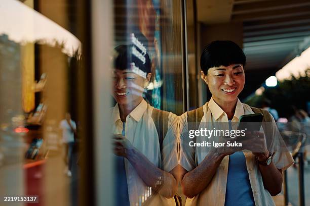 smiling tourist using smartphone in front of a shop window at night - etalage stockfoto's en -beelden