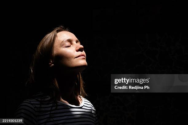 woman enjoying sunlight with peaceful expression in a dark background - schwache beleuchtung stock-fotos und bilder