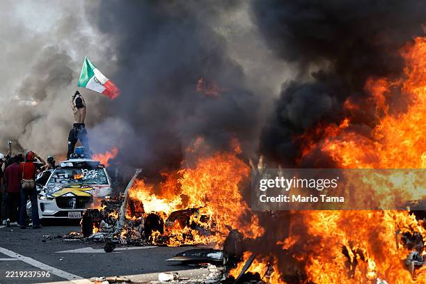 Protestor holds up a Mexican flag as burning cars line the street on June 08, 2025 in Los Angeles, California. Tensions in the city remain high after...