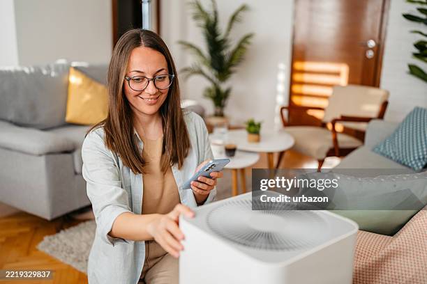 young woman turing on the air purifier using her phone while sitting on the sofa at home - indoor air pollution stock pictures, royalty-free photos & images