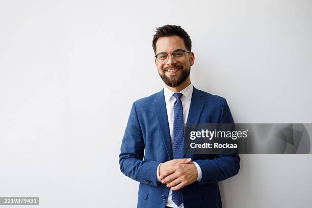 portrait of a cheerful businessman wearing glasses and suit, standing against a white wall with his hands clasped - fato imagens e fotografias de stock